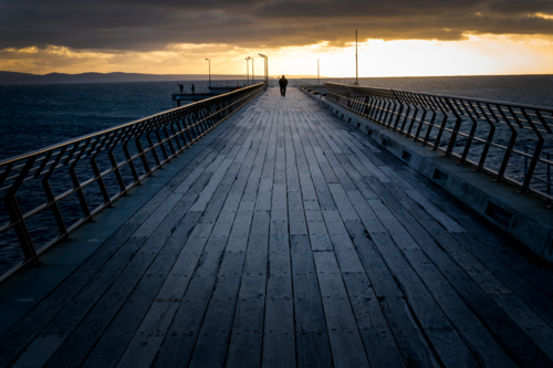 Lone figure walking a weathered pier toward the rising light. - Australian Stock Image