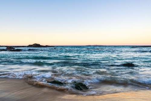 Long exposure of water flowing over rocks with subtle sunset colours reflected in the wet sand. - Australian Stock Image