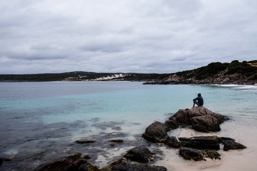 Long shot of man sitting and fishing from rock on remote beach