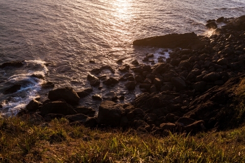 Looking down from a grassy headland as ocean waves crash against large rocks at sunset.