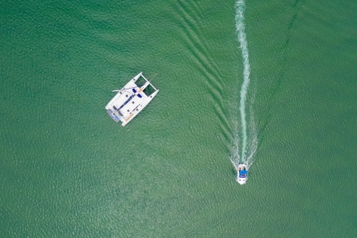 Looking down on a single catamaran and a small boat speeding past leaving a wake in the calm water - Australian Stock Image