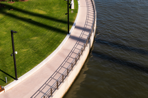 Looking down on curved river bank with path, fence, sharp shadows and green grass lawn - Australian Stock Image