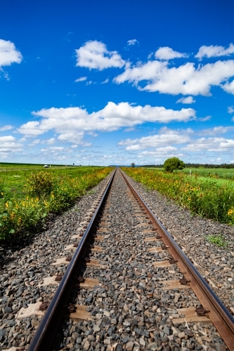 Looking down railway track lined with sunflowers and green to blue sky and clouds above - Australian Stock Image