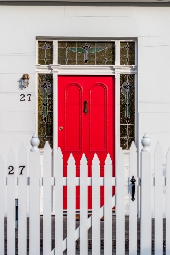 Looking past a picket fence to a bright red entrance door of a white washed sandstone home
