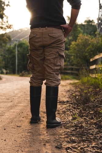 Low angle of man standing on winding dirt road looking at sunset outside rural property - Australian Stock Image