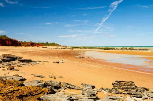 Low tide on an orange sandy beach with red cliffs, rocky foreground and blue sky