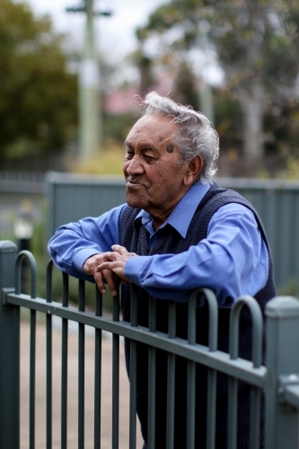 Male Aboriginal elder leaning against fence looking away - Australian Stock Image