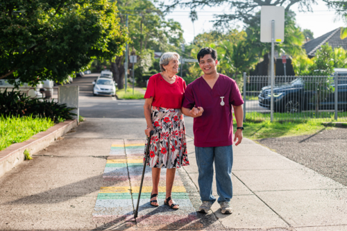 Male caregiver walking with an elderly woman using cane on paved road on sunny day - Australian Stock Image