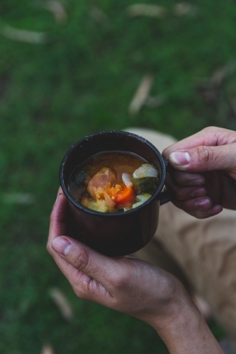 Male hands holding hot mug of soup, sitting on grass on cold afternoon - Australian Stock Image