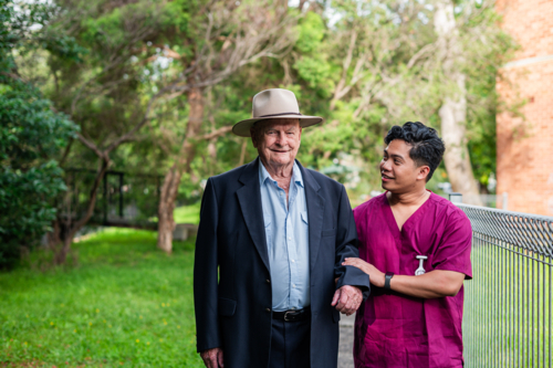 Male healthcare worker assisting an elderly man wearing a suit and wide brimmed hat outdoors - Australian Stock Image
