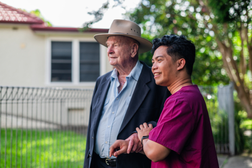 Male healthcare worker assisting an elderly man wearing a suit and wide brimmed hat outdoors - Australian Stock Image