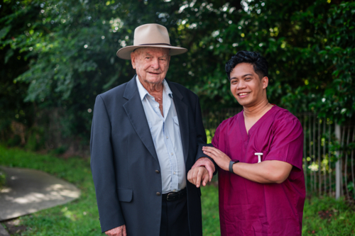 Male healthcare worker assisting an elderly man wearing a suit and wide brimmed hat outdoors - Australian Stock Image
