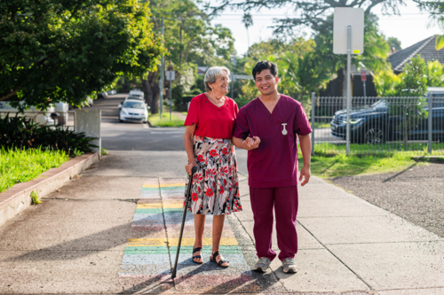 Male healthcare worker assisting an elderly woman walking paved path on sunny day - Australian Stock Image