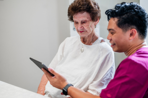 Male healthcare worker holding a tablet device while seated beside an elderly woman - Australian Stock Image
