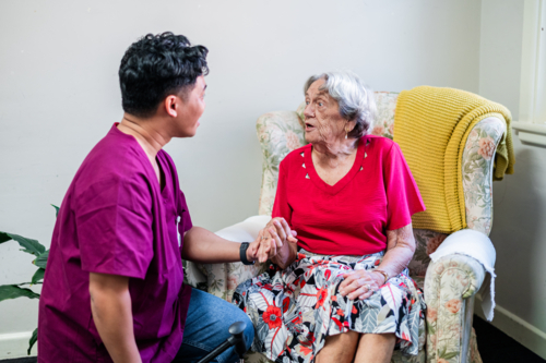 Male healthcare worker holding elderly womans hand seated on patterned armchair in a conversation - Australian Stock Image
