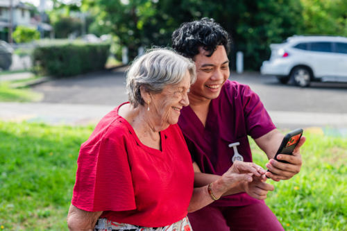 Male healthcare worker holding the phone for an elderly woman while seated on wooden bench - Australian Stock Image