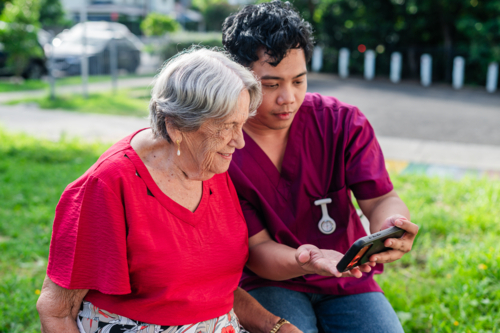 Male healthcare worker holding the phone for elderly woman seated on a bench outdoors on videocall - Australian Stock Image