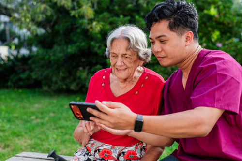 Male healthcare worker holding the phone for elderly woman seated on a bench outdoors on videocall - Australian Stock Image