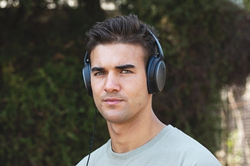 Male in his twenties wearing headphones and listening to music outdoors in the local park - Australian Stock Image