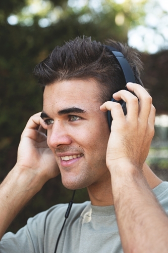 Male in his twenties wearing headphones and listening to music outdoors in the local park - Australian Stock Image