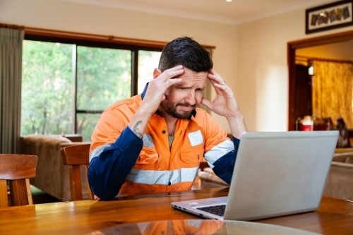 Male tradie in his 30s on laptop feeling overwhelmed and exhausted - Australian Stock Image