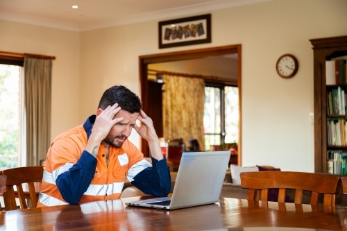 Male tradie in his 30s on laptop feeling overwhelmed and exhausted - Australian Stock Image