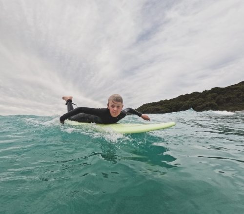 Male youth paddling on soft board in ocean with coastal backdrop on overcast day - Australian Stock Image