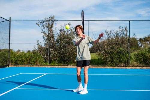 Male youth practices tennis on a hard court with greenery in the background - Australian Stock Image