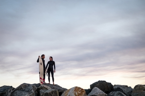 Man and Woman couple standing on coastal rocks holding surfboards looking at each other at sunset