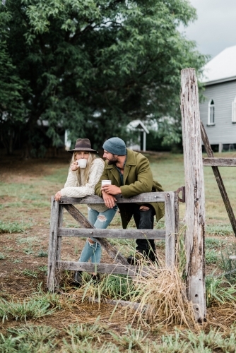 Man and woman leaning on wooden farm gate, drinking coffee