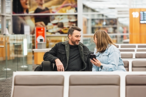 Man and woman watching phone together while waiting in airport departure lounge - Australian Stock Image