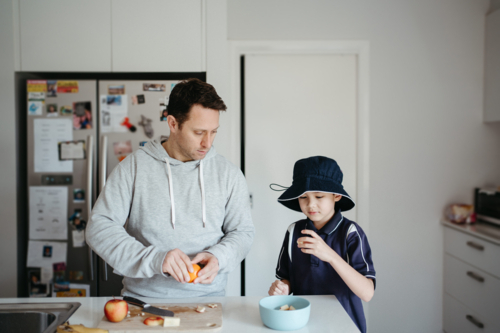 man cutting fruits on the kitchen counter with young boy beside him getting ready for school - Australian Stock Image