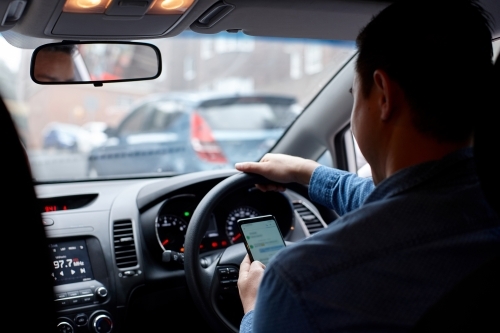 Man driving in his car looking down at mobile phone texting - Australian Stock Image
