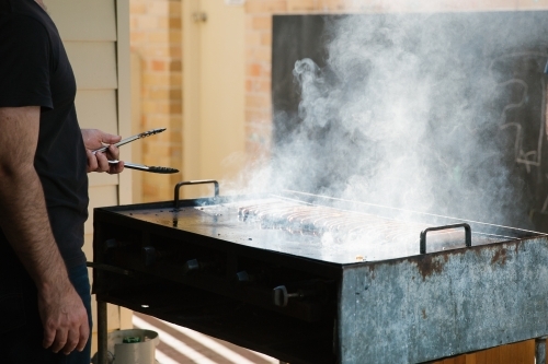 Man grilling and holding a tong in front of a smoking griller
