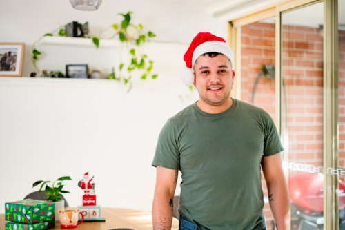 Man in a Santa hat stands happily among festive christmas decorations and wrapped gifts - Australian Stock Image