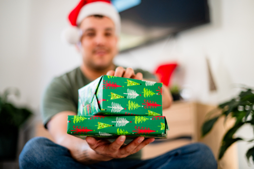 Man in his 30s wearing a Santa hat presents Christmas gifts in a festive setting - Australian Stock Image