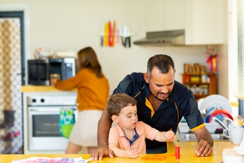man playing with toddler in kitchen with woman in background - Australian Stock Image