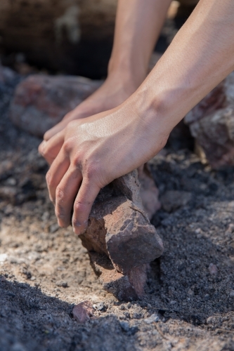 Man preparing camp fire by clearing away ash and charcoal with rock - Australian Stock Image