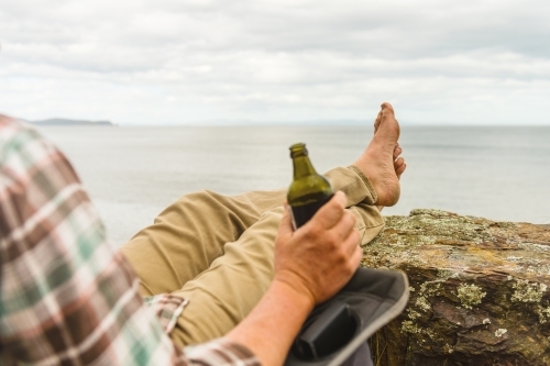 man sitting in tranquil camp spot by the ocean in his chair, with a beer - Australian Stock Image