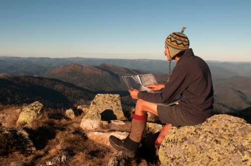 Man sitting on a rock overlooking mountain ranges, looking at a map