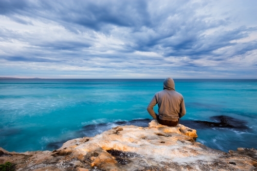 Man sitting on the edge of a rock, looking out at turquoise ocean and cloudy sky - Australian Stock Image
