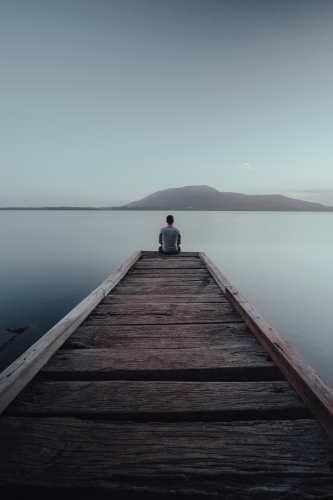 Man sitting on the end of a jetty looking over a lake with a mountain in the background.