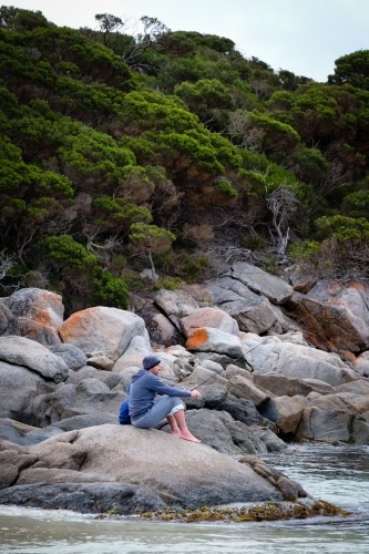 Man sitting with small child behind  and fishing on granite rocks in coastal setting - Australian Stock Image