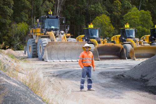 Man standing in the middle of the dirt path with payloaders at the back. - Australian Stock Image