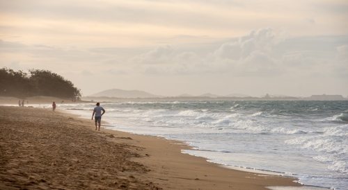 Man walking along Tannum Sands beach with industry in the background, Gladstone Region, Queensland - Australian Stock Image
