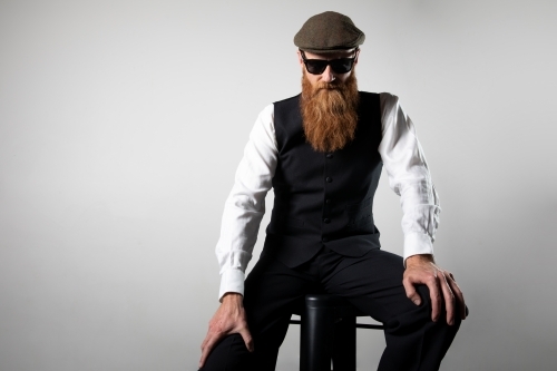 Man with ginger beard and tweed flat cap sitting, posing.