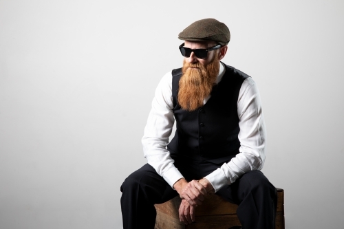 Man with ginger beard and tweed flat cap sitting, posing.