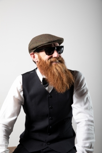 Man with ginger beard and tweed flat cap sitting, posing.