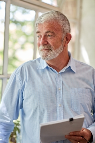 Mature business man holding files while standing, looking our a studio window - Australian Stock Image