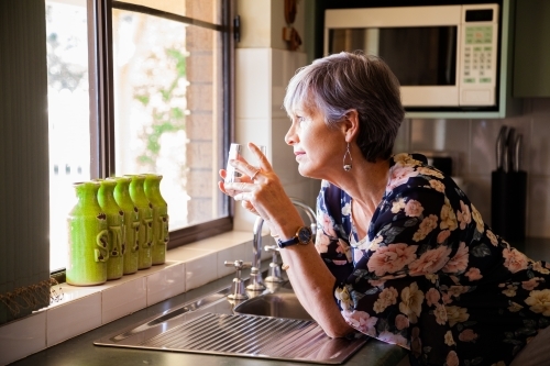 Mature woman holding glass of water kin kitchen looking away - Australian Stock Image